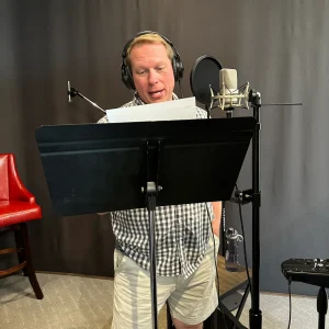C.L. Berns recording narration in a studio while reading from a script into a microphone with headphones on